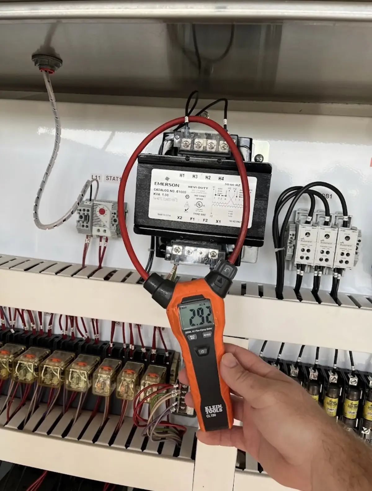 Hand holding a clamp meter reading 2.92 on a conductor inside a control panel with a transformer and contactors